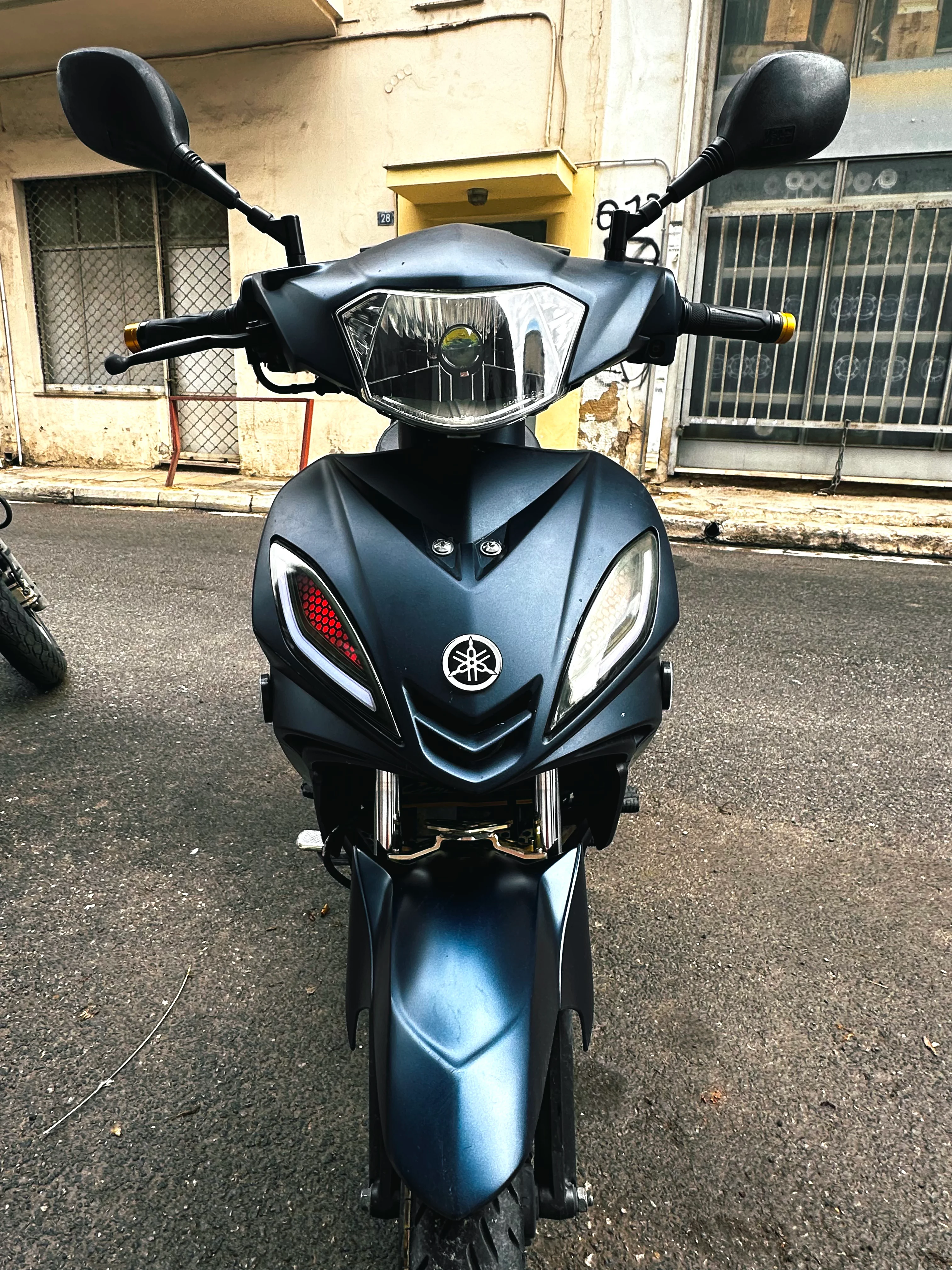 Ματ μπλε σκούτερ Yamaha από μπροστά σε δρόμο, με στρογγυλό φακό προβολέα μέσα στο φανάρι του τιμονιού και φανάρια στην ποδιά, το αριστερό με κόκκινο κυψελωτό ένθετο και λευκή λωρίδα.