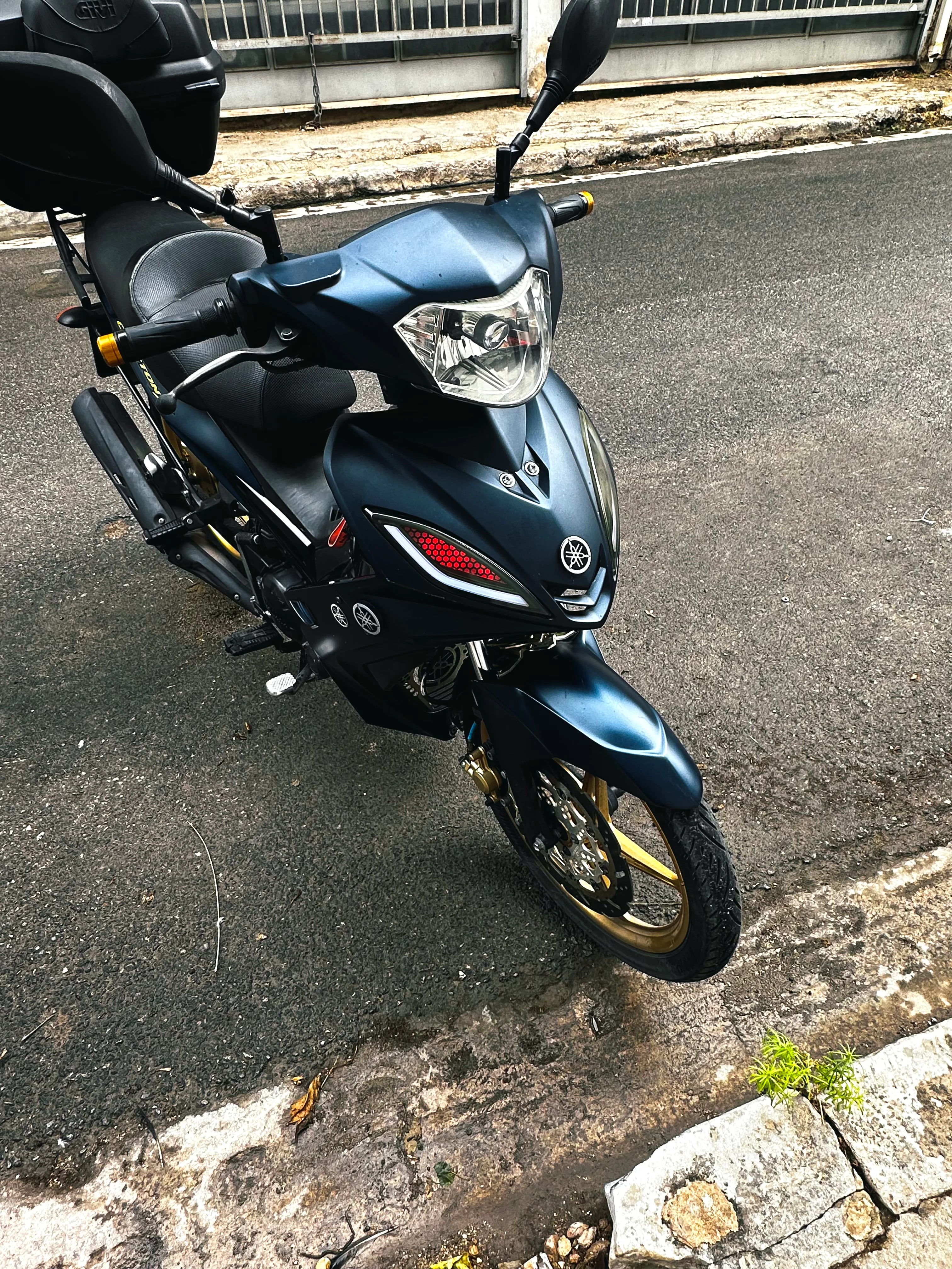 Ματ μπλε σκούτερ Yamaha από μπροστά και λίγο πλάγια, τραβηγμένο από ψηλά σε δρόμο, με στρογγυλό φακό προβολέα μέσα στο φανάρι του τιμονιού, φανάρι με κόκκινο κυψελωτό ένθετο στην ποδιά και χρυσές ζάντες.
